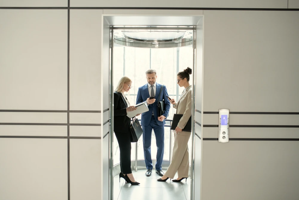 Three business professionals reviewing documents and phones inside a glass elevator.