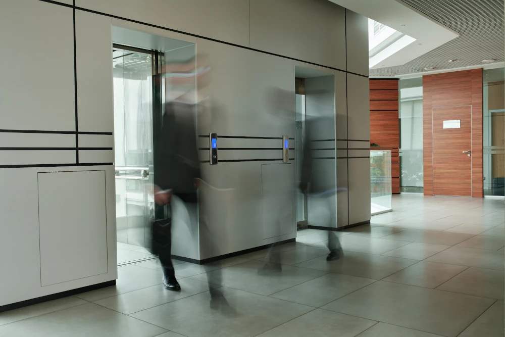 Blurred figures walking past modern glass lift in corporate building lobby.